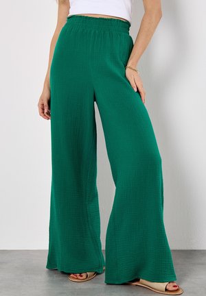 Wide-leg trousers in textured, deep green fabric with an elastic waistband. Paired with beige sandals, showcasing a flowy, casual design.