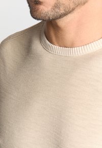 Beige crewneck trui met ribbelhals en een zachte textuur. De stof lijkt zacht en lichtgewicht, geschikt voor casual kleding.