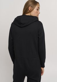 Svart hoodie med avslappnad passform, med dragsko på huven, långa ärmar och ett slätt, texturerat tyg. Bakre vy visar en enkel design.
