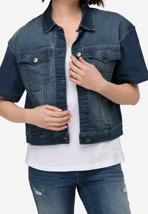 Femme portant une veste en denim foncé à manches courtes sur une chemise blanche et un jean bleu déchiré, ajustant les boutons de la veste.