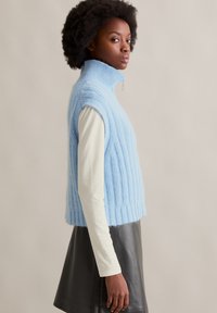 Hellblau gestrickter ärmelloser Rollkragenpullover, gerippte Textur, cropped Länge, kombiniert mit einem cremefarbenen Langarmshirt und einem schwarzen Ledrock.