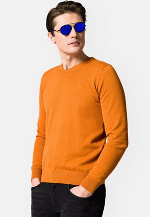Pomarańczowy sweter z dekoltem w serek, z prążkowanymi mankietami i dolnym wykończeniem, noszony na czarnych dżinsach. Model nosi lustrzane niebieskie okulary przeciwsłoneczne. Dyskretne logo na klatce piersiowej.