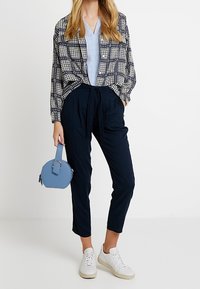 Marinefarbene Hose mit Bindegürtel, hellblaue Bluse, übergroßes schwarz-weiß gemustertes Hemd und runde blaue Handtasche. Weiße Sneakers runden den Look ab.