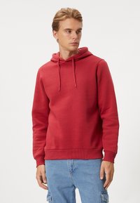 Czerwona bluza z kapturem, wykonana z miękkiego materiału, z kieszenią kangurką, sznurkiem w kapturze oraz prążkowanymi mankietami i dołem. Noszona z jasnoniebieskimi spodniami dżinsowymi.