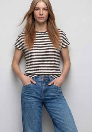 Mujer joven con cabello largo, vestida con una camiseta de manga corta a rayas blanco y negro y jeans azules, de pie con las manos en los bolsillos frente a un fondo liso.