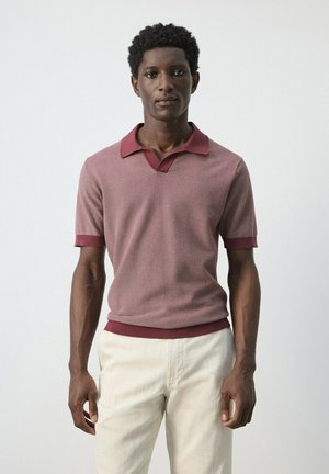 Joven de pie mirando hacia adelante, vistiendo una camisa tipo polo de punto color granate y pantalones beige claro contra un fondo liso y claro.