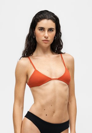Kvinde med langt mørkt krøllet hår iført en rød trekants-bikini top og sorte bikini-bunde mod en ensartet lys baggrund.