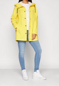 Imperméable jaune avec capuche, boutons-pression à l'avant et poches latérales, porté avec un jean bleu et des baskets montantes blanches.