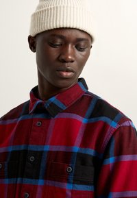 Rode en zwarte flanellen shirt met blauw ruitpatroon, knoopsluiting aan de voorkant, kraag en twee frontzakken. Gemaakt met een crèmekleurige gebreide beanie.