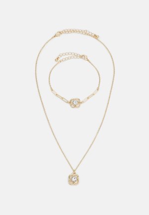 Gouden sieradenset met een halsketting en armband. Beiden hebben een bloemmotief met een centrale heldere edelsteen. De kettingen zijn delicaat en gestructureerd.