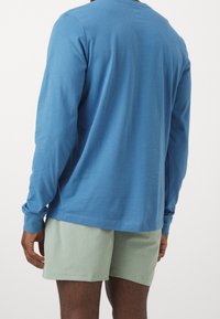 Muž v modré košili s dlouhým rukávem a světlých zelených šortkách, zobrazený zezadu na pozadí jednoduché bílé barvy.