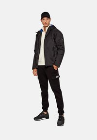 Giacca nera isolante con cappuccio foderato di blu, indossata sopra un maglione chiaro. Abbinata a pantaloni da tuta neri con logo e sneakers nere.