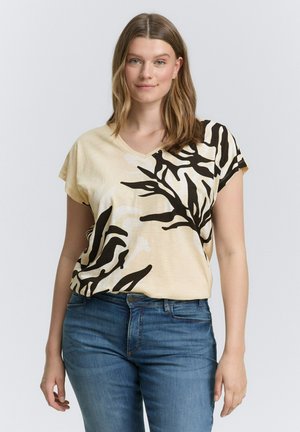 Beige V-neck t-shirt with black abstract leaf print; short sleeves; soft fabric; worn with blue denim jeans.