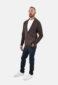 Blazer marrone lavorato a maglia sopra una camicia bianca, jeans blu scuro e sneakers bianche. Il blazer presenta un bottone singolo, rever a lancia e due tasche.