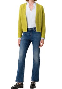 Grüne Strickjacke mit gerippten Kanten, getragen über einer weißen Bluse, kombiniert mit blauen, ausgestellten Jeans und schwarzen Slippern mit silbernen Akzenten.