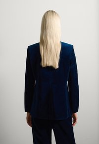 Blazer de terciopelo azul marino con un corte ajustado, mangas largas y una textura suave, combinado con pantalones a juego. Vista posterior.