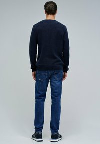 Mann, der mit dem Rücken zur Kamera steht, trägt einen marineblauen Pullover, blaue Jeans mit Gesäßtaschen und schwarze Sneakers vor einem schlichten grauen Hintergrund.