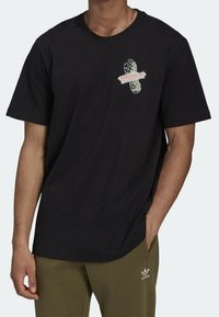 Černé bavlněné tričko s krátkým rukávem, s grafickým designem v bílé a růžové, zobrazující nápis "adidas ADVENTURE" a akcentové vzory.