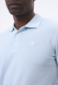 Hellblaues Poloshirt aus strukturiertem Stoff, mit zwei Knöpfen und einem weißen Vogel-Logo auf der Brust. Der Kragen ist umgeschlagen und lässig gestylt.