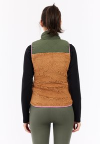 Fleece vest met een groene gewatteerde bovenkant en een bruine gestructureerde onderkant, met een roze afwerking. Draagt over een zwart langemouwen top en groene leggings.
