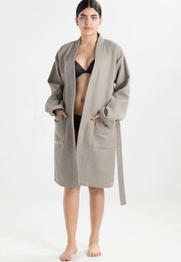 Robe cinzenta texturizada com corte amplo, apresenta mangas largas, bolsos e cintura com cordão. Usada sobre um biquíni preto, descalça contra um fundo branco.