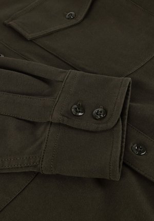 Donkergroene stoffen shirtmouw met twee zwarte knopen op de manchet en een deel van een gesloten borstzak zichtbaar.