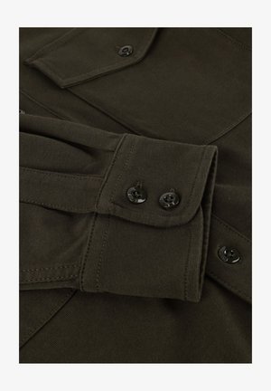 Donkergroene stoffen shirtmouw met twee zwarte knopen op de manchet en een deel van een gesloten borstzak zichtbaar.