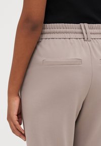 Beige stretchbroek met een soepele textuur, voorzien van twee achterzakken en een verborgen sluiting aan de voorkant. Close-up van de taille en hand.