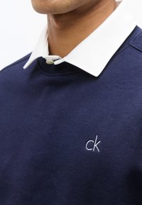 Άνδρας που φοράει μπλε σκούρο φούτερ με λευκό γιακά, με μικρό λευκό κεντητό λογότυπο "ck" στο στήθος.