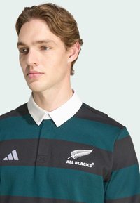T-shirt de rugby à rayures noires et teal avec un col blanc ; présente un logo de fougère brodé et un détail de marque à trois rayures sur la manche.