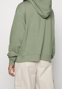 Sweat à capuche vert olive avec capuche à cordon, poignets et ourlet côtelés. Coupe ample et texture douce, associé à un pantalon cargo beige clair.