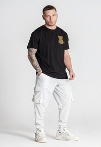 Čierne bavlnené tričko s zlatým logom medveďa, skombinované s bielymi cargo nohavicami s vzorom. Topánky sú biele s textovým detailom na boku.