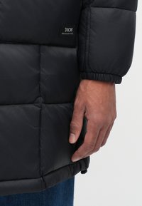 La giacca a vento nera presenta un design trapuntato con polsini elasticizzati e una toppa con il logo. Il tessuto ha una superficie liscia e lucida.