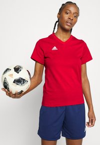 Červené tričko s výstrihom do V a bielym logom Adidas, spárované s tmavomodrými šortkami. Osoba drží vzorovaný futbalový loptu. Jednoduchý dizajn, športový strih.
