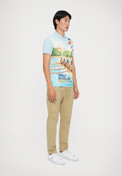 Άνδρας όρθιος με ανοιχτό μπλε πόλο μπλουζάκι με σχέδιο φεστιβάλ κωπηλασίας, μπεζ cargo παντελόνι και λευκά αθλητικά παπούτσια σε αδιάφορο φόντο.