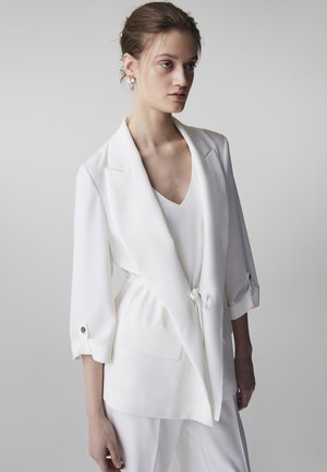 Vrouw draagt een witte blazer met opgerolde mouwen en een ceintuur in de taille over een wit topje, staand tegen een effen neutrale achtergrond.