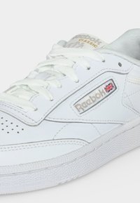 Basket blanche Reebok Classic avec lacets blancs, bout perforé et écusson du logo Reebok avec un drapeau britannique sur le côté.