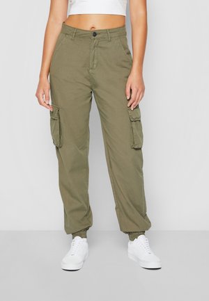 Person iført olivengrønne cargobukser med lommer og hvide sneakers, stående mod en ensfarvet lys baggrund.