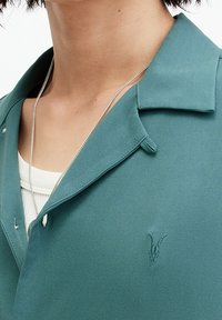 Teal shirt met een zachte stof, een kraag, een enkele knoop aan de voorkant en een geborduurd logo op de borst. Gelaagd over een witte top.