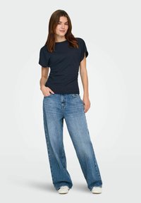 Femme portant un haut noir à manches courtes, un jean bleu ample et des baskets blanches, debout avec une main dans la poche devant un fond uni.