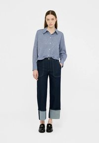 Modrá a biela pruhovaná košeľa na gombíky s golierom, skombinovaná s tmavými džínsami s vysokým pásom a svetlými manžetami z denimu. Čierne mokasíny.