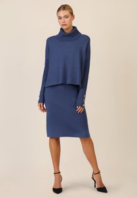 Maglione blu a maglia con colletto alto, abbinato a una gonna a tubino coordinata. Il maglione presenta maniche a costine e dettagli con bottoni ai polsi.