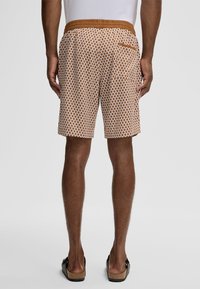 Uomo che indossa pantaloni corti a fantasia marrone e bianco, camicia bianca e sandali neri senza strap, in piedi di spalle su uno sfondo grigio semplice.