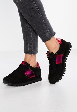 Piernas con jeans negros remangados que llevan zapatillas negras con detalles brillantes en rosa y tachuelas, sobre un fondo de suelo claro.