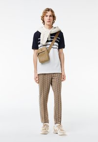 Completo da uomo con polo a righe blu navy, pantaloni beige con fantasia, maglione di maglia crema appoggiato sulle spalle e sneakers beige con dettagli.