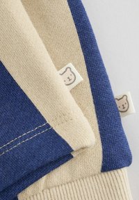 Soft knit fabric pieces in beige and navy blue, each with a small white tag featuring a simple bear face illustration.