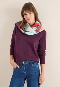 Purple sweater with long sleeves, featuring a rounded hem. Multicolored patterned scarf with geometric designs wrapped around the neck. Denim jeans.