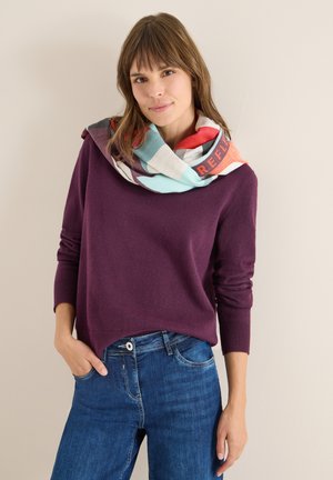 Suéter morado de manga larga con dobladillo redondeado. Bufanda multicolor con diseños geométricos envuelta alrededor del cuello. Vaqueros de mezclilla.