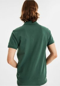 Grön polo med krage, korta ärmar och en strukturerad textil. Bakåtsidan visar en ren, figurnära design utan logotyper.