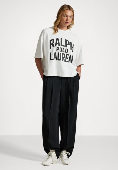 Balts pārmērīga izmēra džemperis ar lielu melnu "Ralph Lauren" logo, apvienots ar brīvām melnām biksēm un gaišas krāsas kedām.